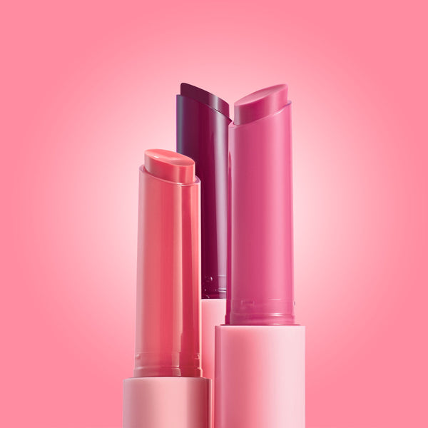Three She’s So Juicy gloss balm sticks stand uncapped, showing coral, berry, and rosy pink bullets against a soft pink gradient, highlighting the ColourPop So Juicy Plumping Gloss Balm with Peptides for shiny, fuller-looking lips.