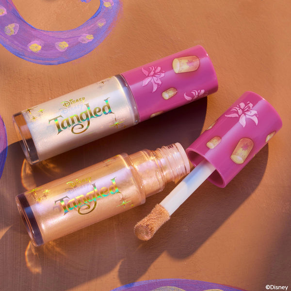 ColourPop Liquid Highlighter in I See the Light, a champagne‑gold shimmer, shown open with a doe‑foot applicator beside a closed tube; Disney Tangled pink caps display floating lanterns and florals on a warm painted backdrop.
