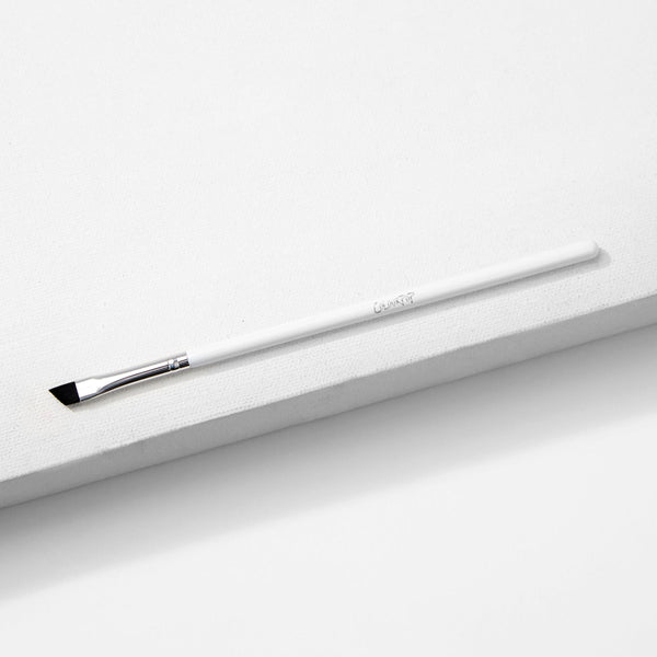 ColourPop Angled Eye Brush resting diagonally on a white textured block, featuring slim angled black bristles, a silver ferrule, and a long white handle - makeup brush for precise eyeliner and brow shaping.