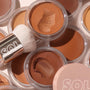 ColourPop SOL Face & Body Bronzing Balm in Dark sits open with a smooth swatch in a clear-lidded jar, surrounded by warm-toned shades and a SOL-branded buffing brush in a flat lay, showcasing the creamy texture and sun‑kissed finish.