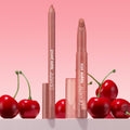 Brink ColourPop Lippie Pencil and Lippie Stix stand upright with caps off, revealing warm rosy nude tips, arranged among glossy cherries on a white surface against a pink gradient background.