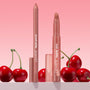 Brink ColourPop Lippie Pencil and Lippie Stix stand upright with caps off, revealing warm rosy nude tips, arranged among glossy cherries on a white surface against a pink gradient background.