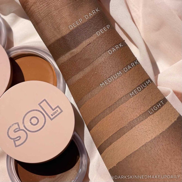 ColourPop SOL Face & Body Bronzing Balm in shade Dark swatched on a forearm, with labeled stripes Light, Medium, Medium Dark, Dark, Deep, Deep Dark; open SOL Body tins nearby on beige fabric, showing the creamy bronze finish and shade range.