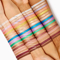 Friday Feels Shadow Stix swatch - one creamy, shimmery stripe - glides across multiple forearms of different skin tones, shown alongside the full ColourPop Shadow Stix lineup to demonstrate pigment payoff and blend in bright studio lighting.