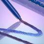ColourPop Chrome Crème Gel Liner Ventura Blvd draws a shimmering blue‑purple chrome line on a glossy surface, the blue pencil barrel with celestial icons shown close up against a pastel lilac gradient studio backdrop.