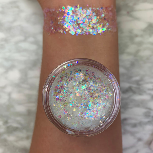 Disco Lady glitter gel swatched across a forearm, displaying chunky iridescent rainbow sparkle; an open pot of translucent gel packed with holographic flakes rests on the arm over a marble surface, showcasing payoff and texture; ColourPop Glitter Gel.