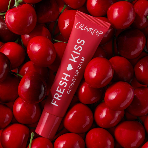 ColourPop Fresh Kiss Glossy Lip Balm in Cherry Glaze rests among glossy red cherries in a top-down flatlay, showcasing the Fruit Basket Glossy Lip Balm Trio - ColourPop Glossy Lip Balm Trio offering hydrating, high-shine cherry tint.