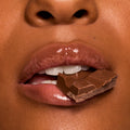 Glossy lips bite a piece of milk chocolate, showcasing rich shine in a chocolate-brown shade from Gloss Goals, a ColourPop Full Collection Set, in a tight beauty macro emphasizing a smooth, hydrated finish.