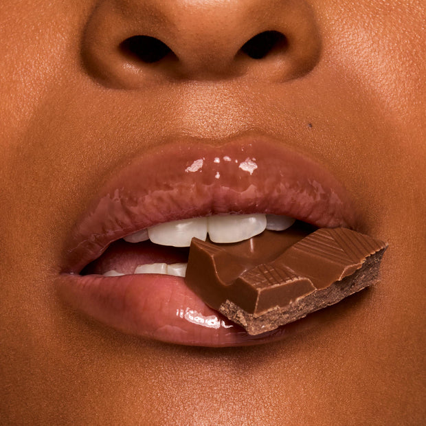 Glossy lips bite a piece of milk chocolate, showcasing rich shine in a chocolate-brown shade from Gloss Goals, a ColourPop Full Collection Set, in a tight beauty macro emphasizing a smooth, hydrated finish.