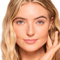 Model wearing ColourPop Fresh Kiss Glossy Balm in Strawberry Milk, showcasing a sheer pink glossy lip finish; full-face studio close-up with natural makeup, soft waves, and a hand at her cheek against a white background.