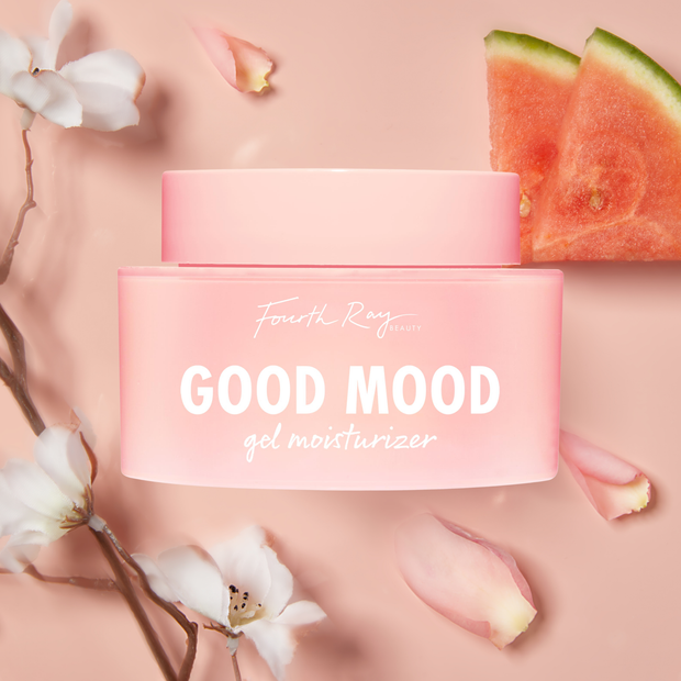 Good Mood Gel Moisturizer jar by Fourth Ray Beauty sits centered on a blush-pink backdrop, the pink frosted container with white lettering surrounded by white blossoms, petals, and watermelon slices - styled ColourPop Face Moisturizer product shot.