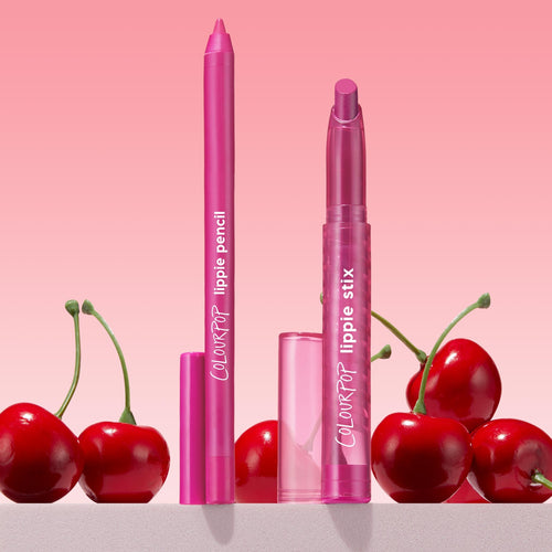 Heart Lips ColourPop Lippie Stix + Pencil Set stands upright with caps off, revealing hot-pink lipstick and matching liner in translucent pink tubes labeled ColourPop, styled among glossy red cherries on a pink gradient background.