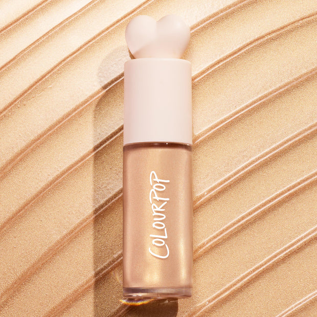 ColourPop Liquid Highlighter in Sugar Donut stands upright with a heart-shaped cap, showcasing a luminous champagne‑gold glow in a frosted tube, set against sweeping metallic swatches that emphasize its shimmery finish.