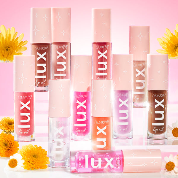 ColourPop Lux Lip Oil bottles from the Comb By collection stand in rows, peach caps with white star icons and bold LUX lettering, against a rosy backdrop with daisies, showing clear, coral, pink, mauve, and deep brown shades for glossy, hydrating lips.