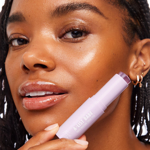 A-List Babe Serum Stick held near a model’s cheek, the lavender twist-up ColourPop Serum Stick showing a sheer purple balm tip; close-up highlights glossy lips and radiant, dewy skin in a studio beauty shot.
