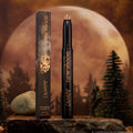 Vampire Girl ColourPop Shadow Stix stands uncapped, showing a pink‑green multichrome tip on a sleek black tube with gold lettering, its box beside it, set on a rocky surface with pine trees and a glowing full moon background.