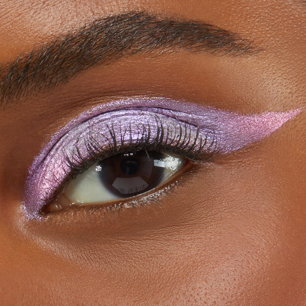 Close-up of an eye wearing Colourpop Cotton Candy Shadow Stix, showing shimmering pastel pink and lavender cream eyeshadow blended across the lid and extended into a sharp wing on smooth medium-brown skin.