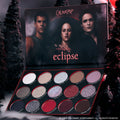 ColourPop Eclipse shadow palette from the Twilight collection lying open in a dark forest setting, showing 12 shimmer and matte cool-toned and red eyeshadows arranged in rows beneath moody character artwork on the inner lid.