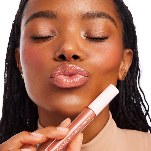 Glossed lips pout as a model holds the ColourPop It Girl Ultra Glossy Lip tube near her mouth, showcasing a high-shine, sheer nude shimmer finish in a clean white studio close-up.