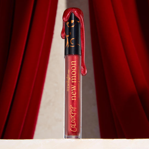 Runs with Vampires Ultra Glossy Lip tube standing upright with metallic blood‑red gloss dripping over a black cap, framed by rich red velvet curtains on a stone ledge, a ColourPop Ultra Glossy Lip delivering high‑shine, vampy color.