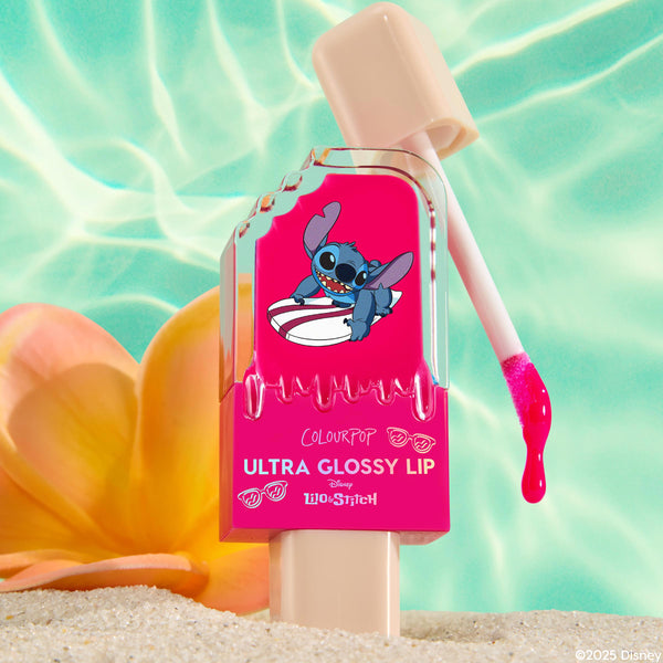 ColourPop Ultra Glossy Lip Surfer Vibes tube stands in beach sand as hot-pink gloss drips from the doe-foot applicator, featuring Stitch on a surfboard on the bottle, set before a plumeria flower and shimmering turquoise pool backdrop.