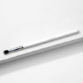 ColourPop Small Tapered Brush resting diagonally on a white textured platform, featuring a slim white handle, chrome ferrule, and small tapered black-and-white bristles for precise eyeshadow blending and detail work.