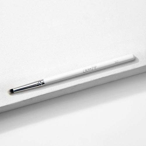 ColourPop Small Pencil Brush makeup brush lying diagonally on a white textured block, showing a slim white handle, silver ferrule, and small tapered bristles for precise eyeshadow application and smudging.