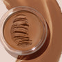 Open glass jar of ColourPop SOL Face & Body Bronzing Balm in Dark, its creamy bronzer surface swiped into ridges, set on a backdrop of blended warm-brown pigment.