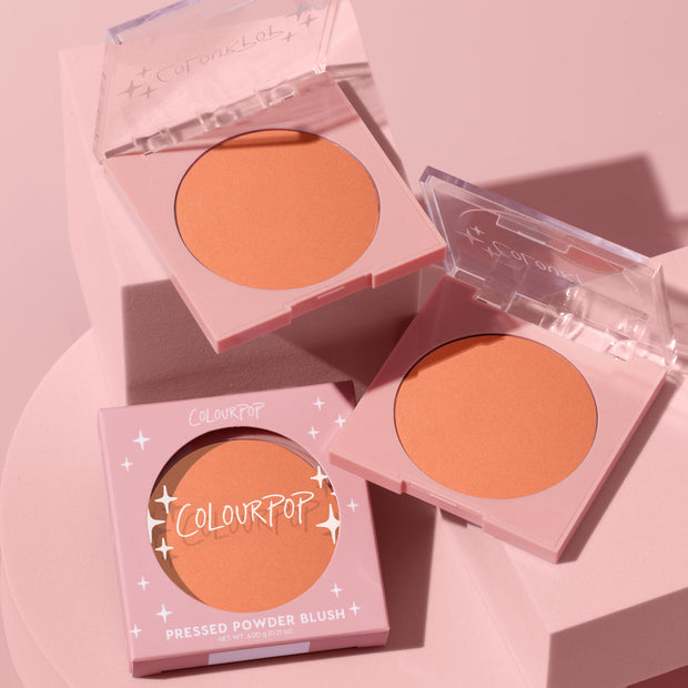 ColourPop Pressed Powder Blush Pretty Toasty compacts sit open on a pink pedestal, revealing a warm peachy-terracotta pan, with one boxed unit in blush-pink packaging with star accents and clear lids showcasing the shade.