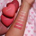 Let’s Dance pressed powder blush swatch, a soft rosy matte, shown on a forearm beside Kiss N Tell, Flirt Alert, 4ever Yours, Text Me, and Babycakes, with heart-shaped ColourPop compacts on pink heart fabric; ColourPop Pressed Powder Blush.